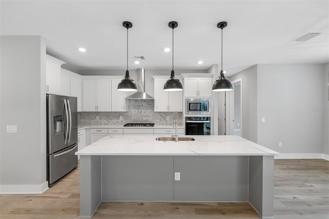 kitchen with wall chimney range hood, stainless steel appliances, pendant lighting, white cabinets, and a kitchen island with sink