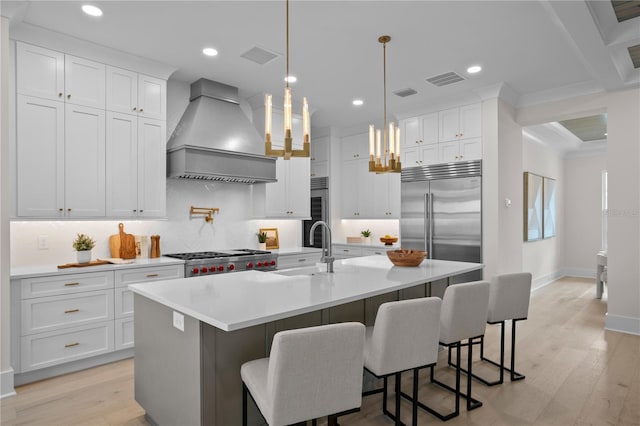 kitchen featuring premium range hood, an island with sink, a kitchen bar, hanging light fixtures, and stainless steel built in fridge