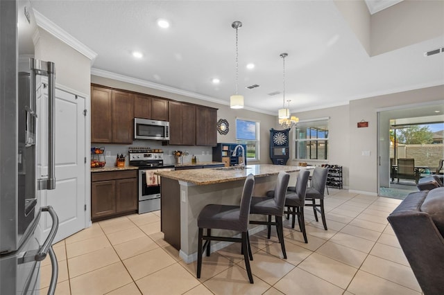 kitchen featuring a center island with sink, light stone counters, stainless steel appliances, crown molding, and decorative light fixtures