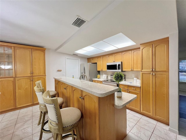 kitchen featuring stainless steel appliances, light countertops, glass insert cabinets, a kitchen island with sink, and a kitchen breakfast bar