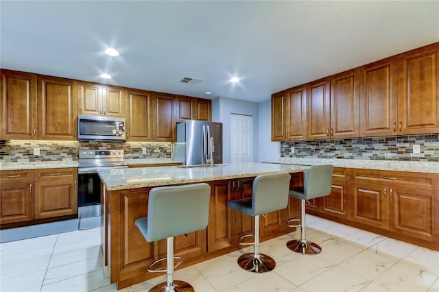 kitchen with backsplash, stainless steel appliances, light stone counters, and a kitchen island with sink