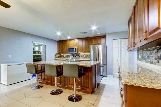 kitchen featuring a center island with sink, light stone countertops, tasteful backsplash, a kitchen bar, and stainless steel appliances