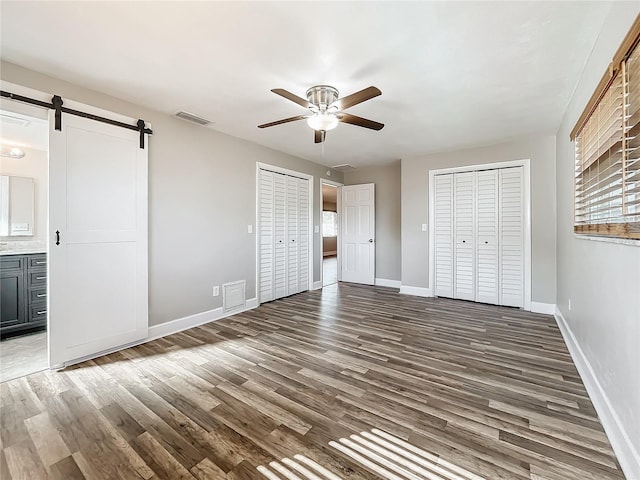 unfurnished bedroom featuring two closets, ceiling fan, a barn door, connected bathroom, and dark hardwood / wood-style floors