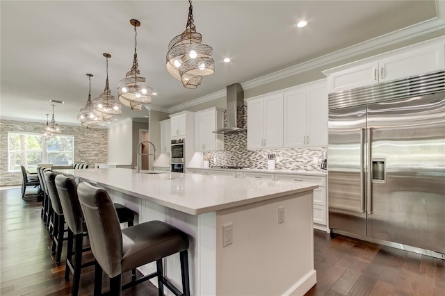 kitchen featuring pendant lighting, a large island with sink, white cabinets, stainless steel built in fridge, and wall chimney range hood