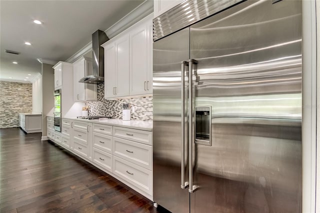 kitchen with appliances with stainless steel finishes, wall chimney exhaust hood, dark wood-type flooring, crown molding, and white cabinets