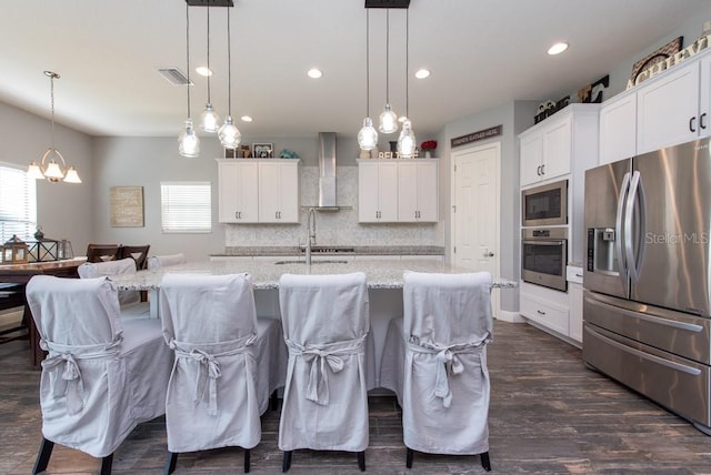 kitchen with an island with sink, wall chimney exhaust hood, hanging light fixtures, and appliances with stainless steel finishes