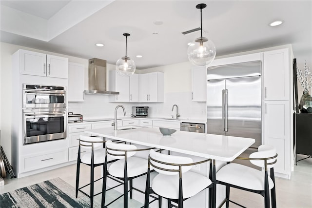 kitchen with sink, an island with sink, stainless steel appliances, wall chimney exhaust hood, and pendant lighting