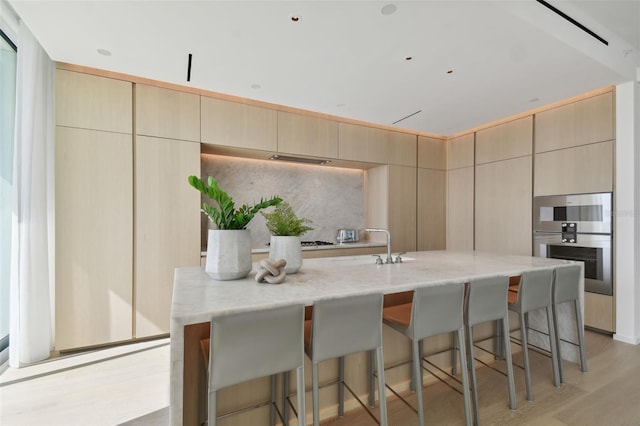 kitchen with light wood-type flooring, light brown cabinets, light stone countertops, and a kitchen island with sink