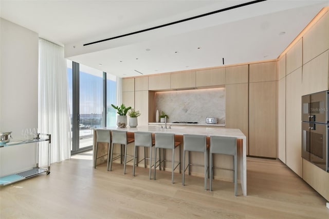 kitchen featuring a kitchen bar, floor to ceiling windows, an island with sink, backsplash, and light wood-type flooring