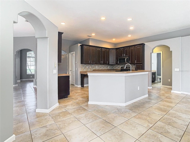 kitchen with appliances with stainless steel finishes, an island with sink, dark brown cabinets, decorative backsplash, and light tile patterned floors