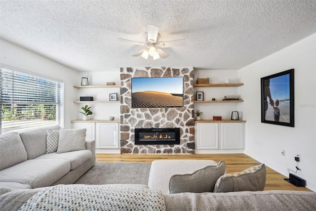 living room with ceiling fan, light hardwood / wood-style floors, a stone fireplace, and a textured ceiling