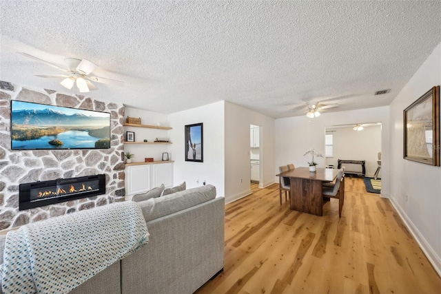 living room featuring a stone fireplace, light hardwood / wood-style floors, a textured ceiling, and ceiling fan