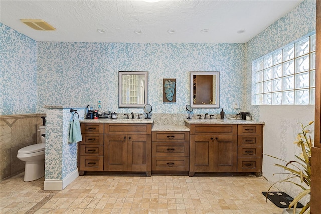 full bathroom with a sink, toilet, double vanity, and wallpapered walls