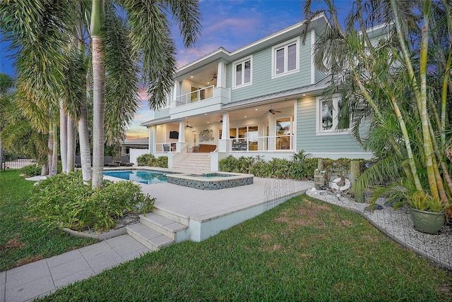 back house at dusk with a yard, a swimming pool with hot tub, a balcony, and ceiling fan