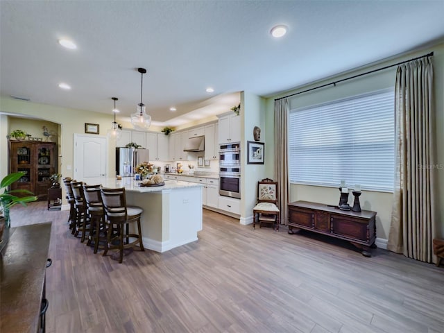 kitchen featuring stainless steel appliances, white cabinets, an island with sink, pendant lighting, and light wood-type flooring