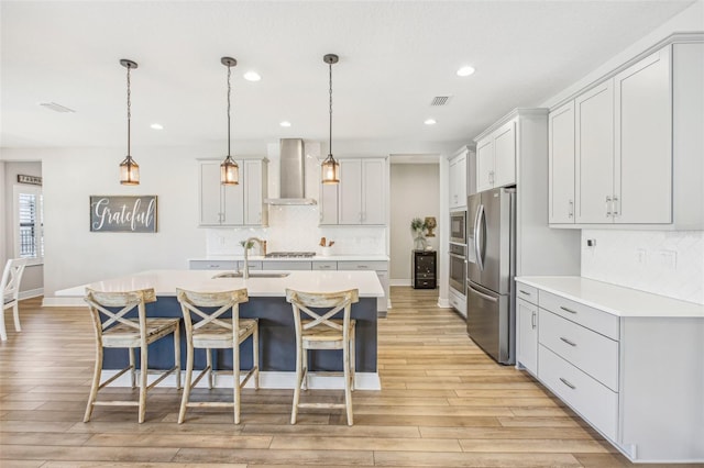 kitchen with decorative light fixtures, wall chimney exhaust hood, stainless steel appliances, and a kitchen island with sink