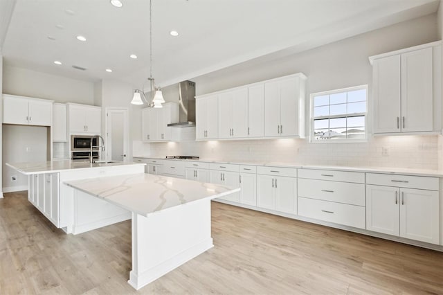 kitchen featuring a kitchen island with sink, white cabinets, wall chimney exhaust hood, stainless steel microwave, and pendant lighting