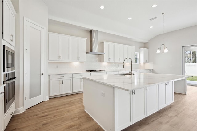 kitchen featuring a sink, white cabinets, wall chimney exhaust hood, a center island with sink, and pendant lighting