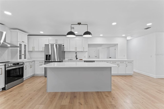 kitchen featuring a center island, wall chimney range hood, appliances with stainless steel finishes, decorative light fixtures, and light hardwood / wood-style flooring