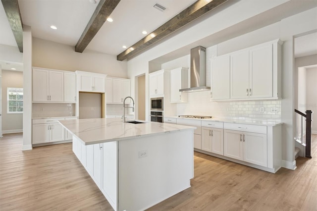 kitchen with wall chimney exhaust hood, appliances with stainless steel finishes, a center island with sink, and white cabinets