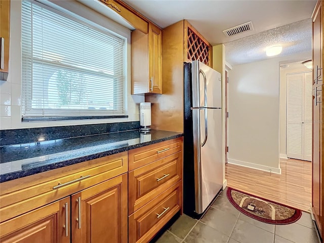 kitchen with light tile patterned flooring, dark stone counters, backsplash, a textured ceiling, and stainless steel refrigerator