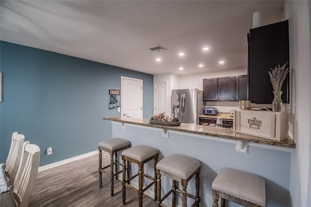 kitchen featuring stainless steel fridge, dark brown cabinetry, stone countertops, hardwood / wood-style flooring, and a kitchen breakfast bar