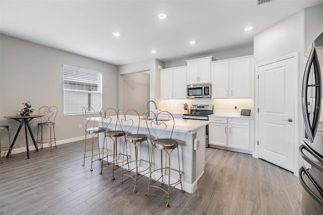 kitchen with a center island with sink, stainless steel appliances, light wood-type flooring, white cabinetry, and a kitchen bar