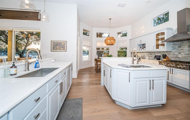 kitchen featuring wall chimney exhaust hood, sink, decorative light fixtures, a kitchen island with sink, and white cabinets