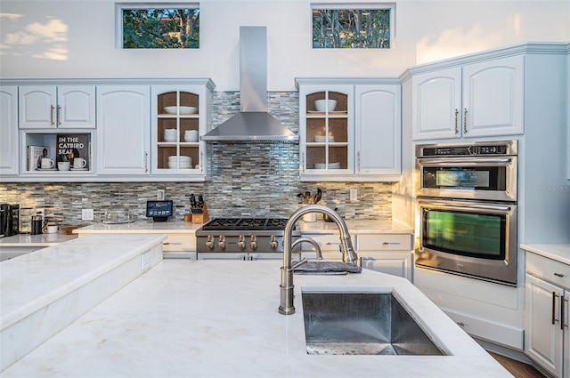 kitchen with sink, wall chimney exhaust hood, tasteful backsplash, light stone countertops, and stainless steel double oven