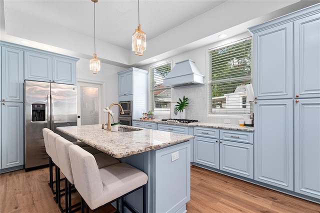 kitchen featuring hanging light fixtures, a kitchen island with sink, light wood-type flooring, appliances with stainless steel finishes, and premium range hood