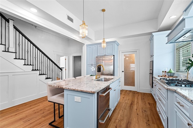 kitchen featuring light hardwood / wood-style floors, a center island with sink, range hood, appliances with stainless steel finishes, and decorative light fixtures