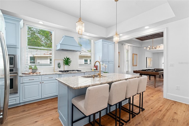 kitchen with appliances with stainless steel finishes, sink, light hardwood / wood-style floors, a tray ceiling, and a kitchen island with sink