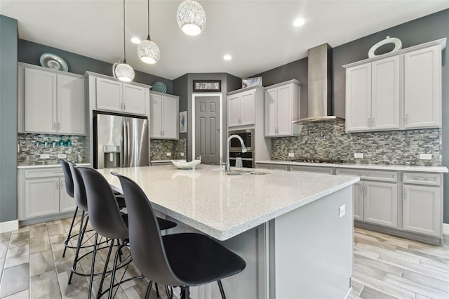 kitchen featuring pendant lighting, appliances with stainless steel finishes, wall chimney range hood, and an island with sink