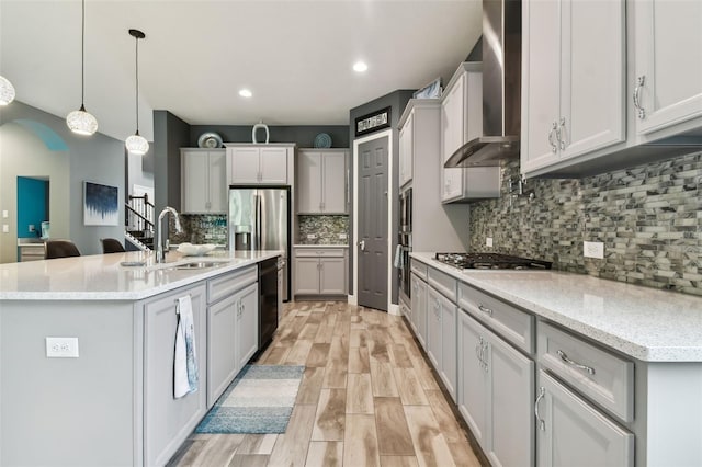 kitchen with a center island with sink, appliances with stainless steel finishes, pendant lighting, sink, and wall chimney exhaust hood