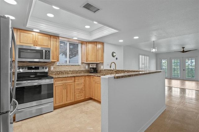 kitchen with kitchen peninsula, appliances with stainless steel finishes, stone countertops, decorative backsplash, and a tray ceiling