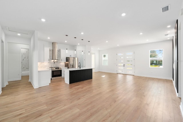 kitchen featuring appliances with stainless steel finishes, an island with sink, wall chimney exhaust hood, white cabinetry, and decorative light fixtures