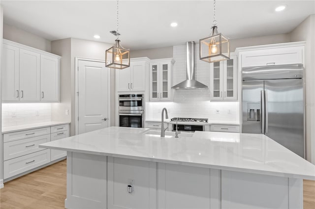 kitchen with appliances with stainless steel finishes, wall chimney exhaust hood, a center island with sink, white cabinets, and hanging light fixtures