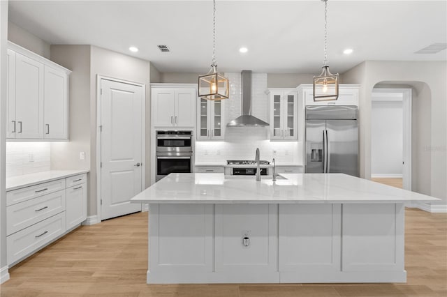 kitchen with wall chimney exhaust hood, white cabinetry, an island with sink, and appliances with stainless steel finishes