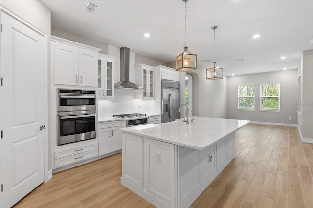 kitchen featuring appliances with stainless steel finishes, wall chimney exhaust hood, white cabinetry, hanging light fixtures, and an island with sink