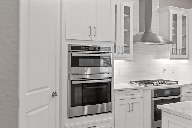 kitchen featuring white cabinets, wall chimney exhaust hood, light stone counters, and appliances with stainless steel finishes