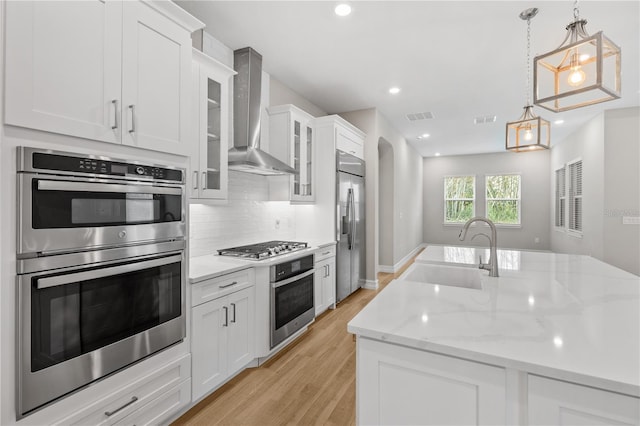 kitchen with white cabinetry, sink, hanging light fixtures, stainless steel appliances, and wall chimney range hood