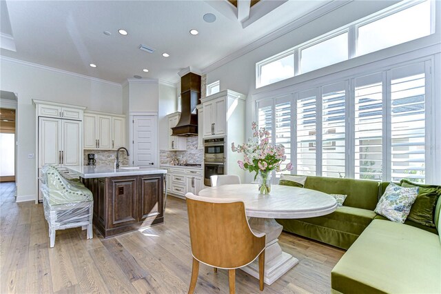 kitchen with light hardwood / wood-style flooring, stainless steel double oven, an island with sink, a kitchen bar, and custom range hood