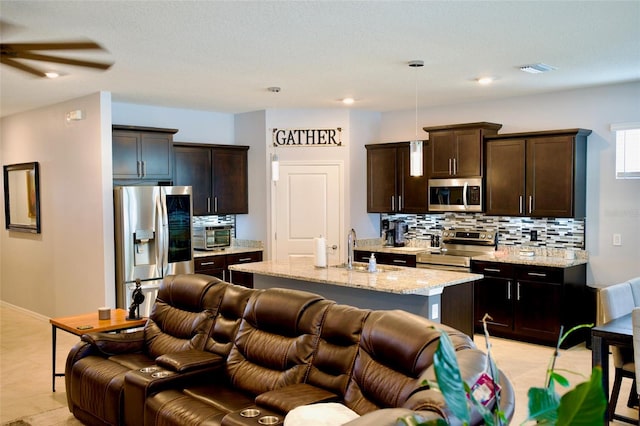 kitchen featuring light stone counters, appliances with stainless steel finishes, an island with sink, dark brown cabinets, and pendant lighting