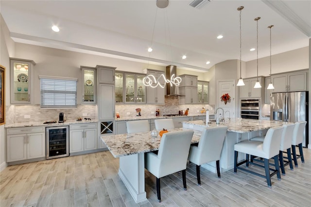 kitchen with a large island with sink, appliances with stainless steel finishes, decorative light fixtures, beverage cooler, and wall chimney range hood