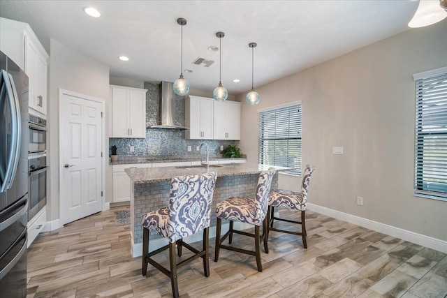 kitchen featuring an island with sink, wall chimney range hood, white cabinetry, and stainless steel appliances