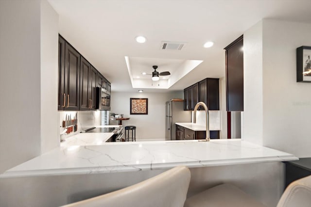 kitchen featuring ceiling fan, stainless steel appliances, a raised ceiling, light stone counters, and kitchen peninsula