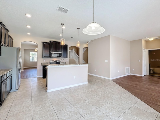 kitchen with stainless steel appliances, dark brown cabinetry, light wood-type flooring, an island with sink, and pendant lighting