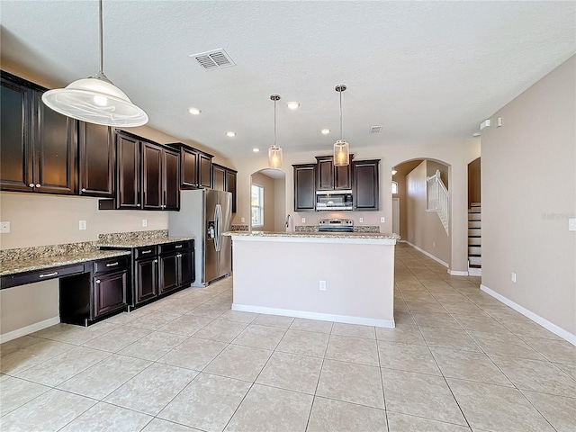 kitchen featuring stainless steel appliances, dark brown cabinets, pendant lighting, light stone countertops, and a kitchen island with sink