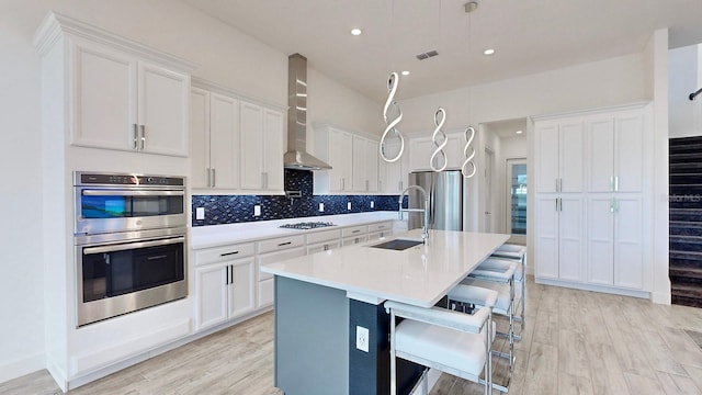 kitchen with sink, wall chimney exhaust hood, an island with sink, white cabinets, and appliances with stainless steel finishes