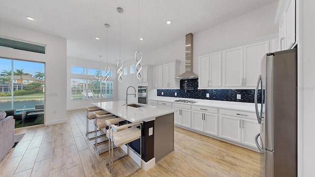 kitchen featuring pendant lighting, wall chimney range hood, an island with sink, white cabinetry, and stainless steel appliances